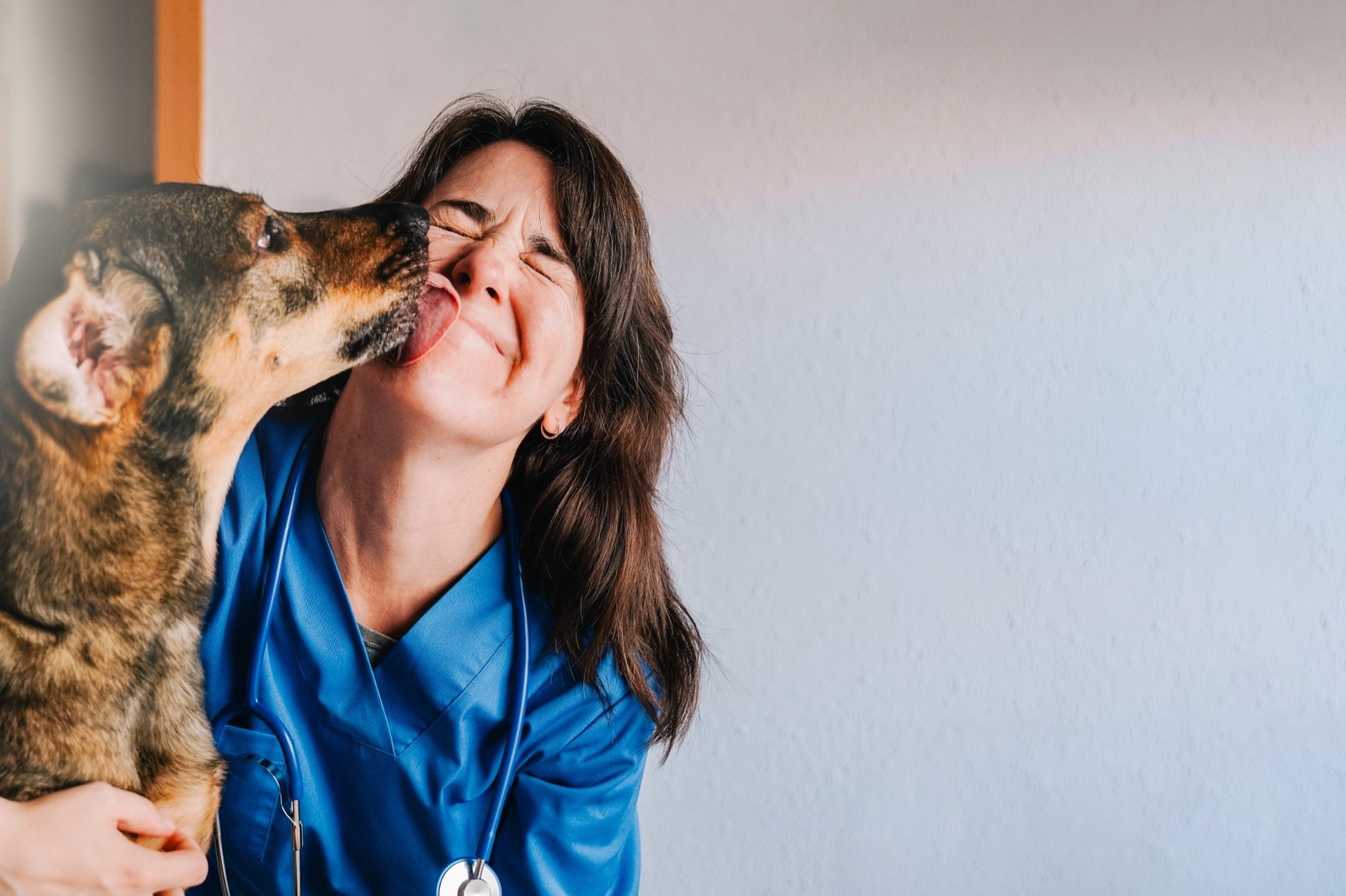 A vet in blue scrubs, eyes shut, as a dog licks her cheek