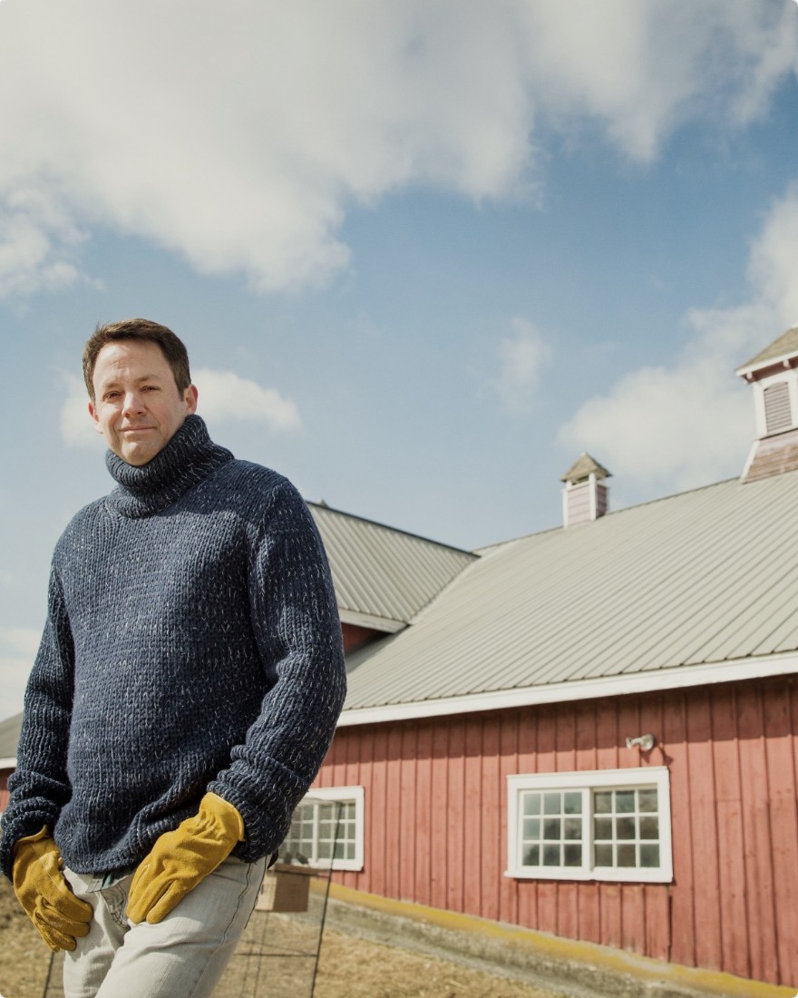 A farmer in a sweater and work gloves in front of a red barn